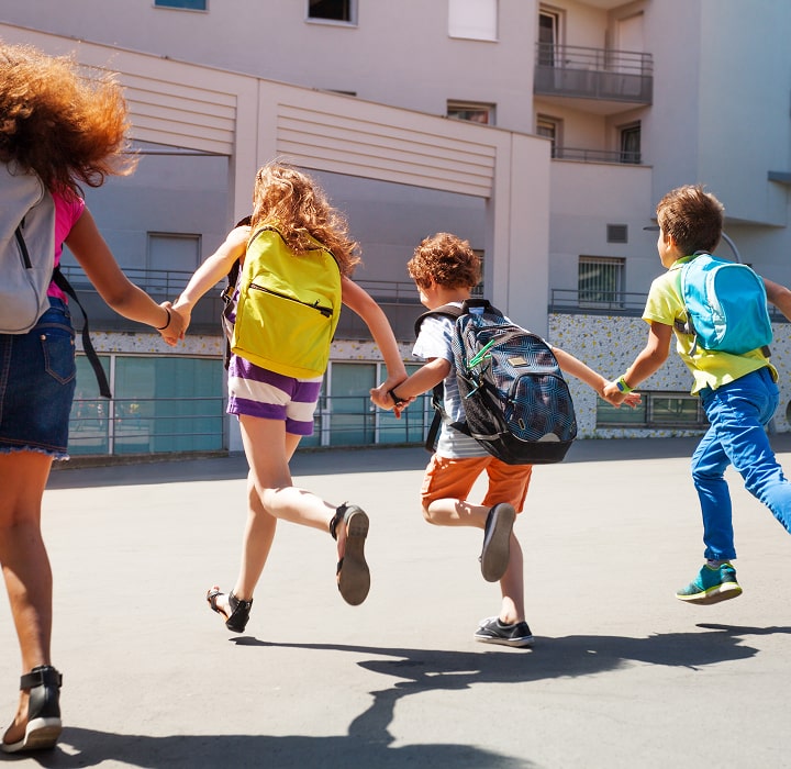 Four children with backpacks running hand in hand toward a school building.