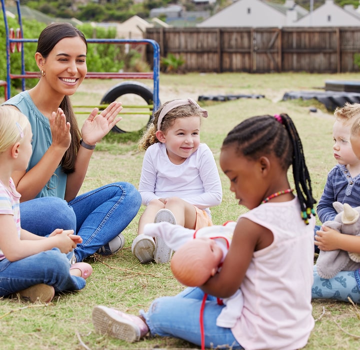 Woman and children sitting in a circle on grass, clapping hands.
